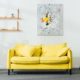 Ein Zimmer mit einem gelben Sofa, einer Lampe, Pflanzen sowie einem Bild und einem Regal an der Wand | Home Staging