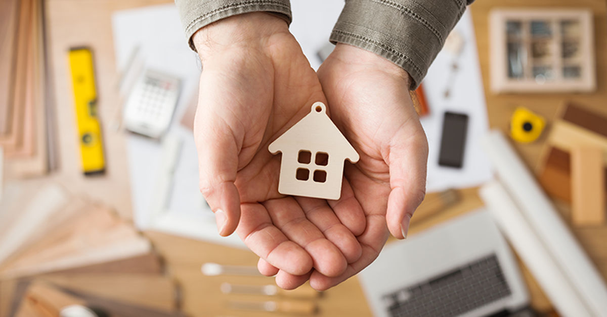 Real estate agent holding a small house, desktop with tools, wood swatches and computer on background, top view; Gebäudeenergie